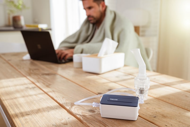 Mann sitzt vor Laptop am Tisch und arbeitet, er hat eine Decke um die Schultern, neben ihm steht eine Taschentücherbox und der Beurer IH 15 Inhalator auf dem Tisch