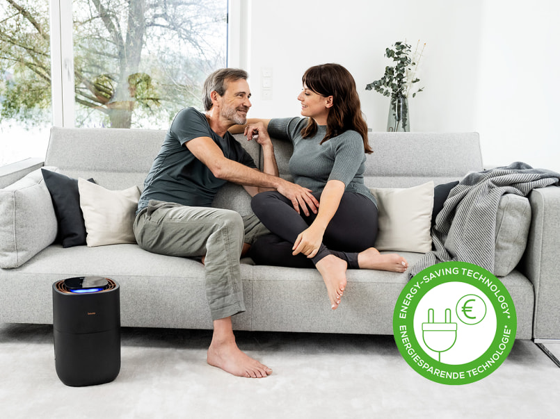 Ein Mann und eine Frau sitzen auf einem grauen Sofa und lächeln sich an. Der LB 300 steht links vom Sofa und leuchtet.