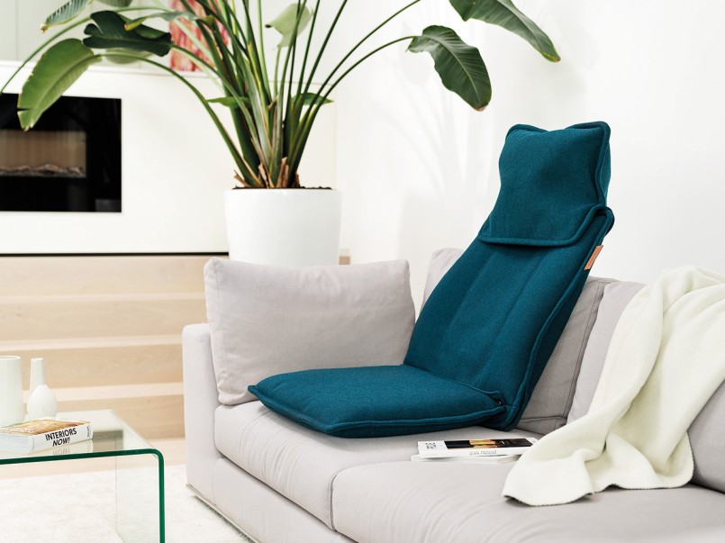 Die Massageauflage MG 330 in petrol liegt als Sitz auf dem Sofa, das in einem modern eingerichteten Wohnzimmer mit Pflanzen steht