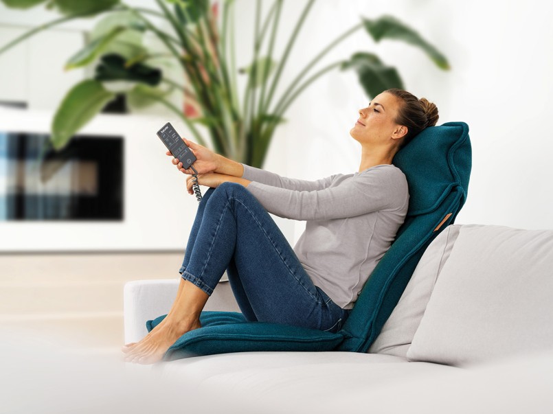 Junge Frau sitzt mit angewinkelten Beinen auf der Massageauflage, hält die Bedienung des Geräts in der Hand und genießt