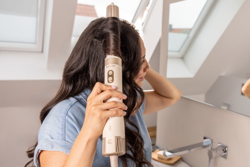Femme aux cheveux foncés se tient devant le miroir et se fait des boucles avec l’embout fer à friser du multistyler