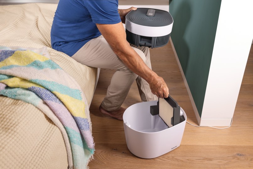 Ein älterer Mann sitzt auf einem Bett und hält die Abdeckung des maremed® Meeresklimagerät in der linken Hand. Der Filter des Meeresklimageräts ist zu sehen.