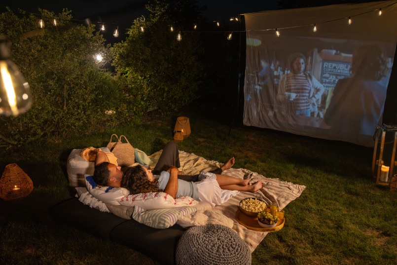 Mann und Frau liegen eingekuschelt auf einer Decke mit vielen Kissen draußen und schauen einen Film auf einer Leinwand, es ist dunkel, neben ihnen steht Popcorn und Trauben
