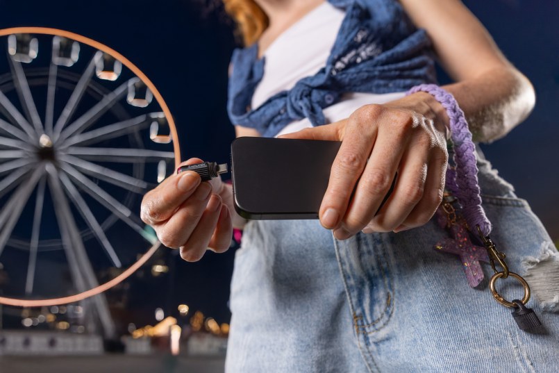 Frau steht bei Nacht draußen, im Hintergrund ist ein beleuchtetes Riesenrad. Man sieht sie nur ab dem Hals abwärts. Sie hält in der Hand ein Handy und steckt darein den Beurer BiteX Go Insektenstichheiler.