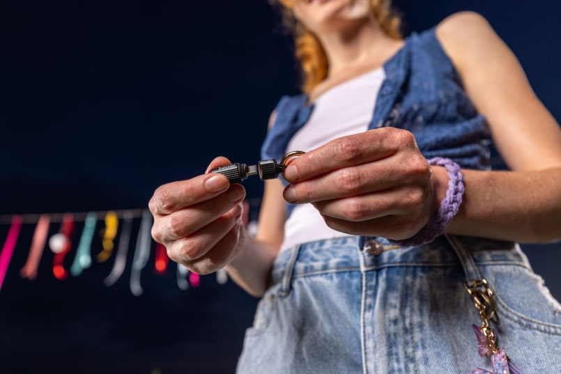 Frau steht bei Nacht draußen, im Hintergrund ist eine bunte Girlande. Man sieht sie nur ab dem Hals abwärts. Sie hält in der Hand den Beurer BiteX Go Insektenstichheiler, in einer Hand hält sie den Deckel, in der anderen das Gerät.