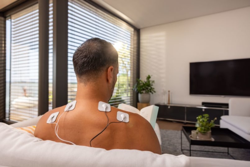 Man sitzt auf dem Sofa zum Rücken und er hat an seinem Rücken Elektroden angebracht.