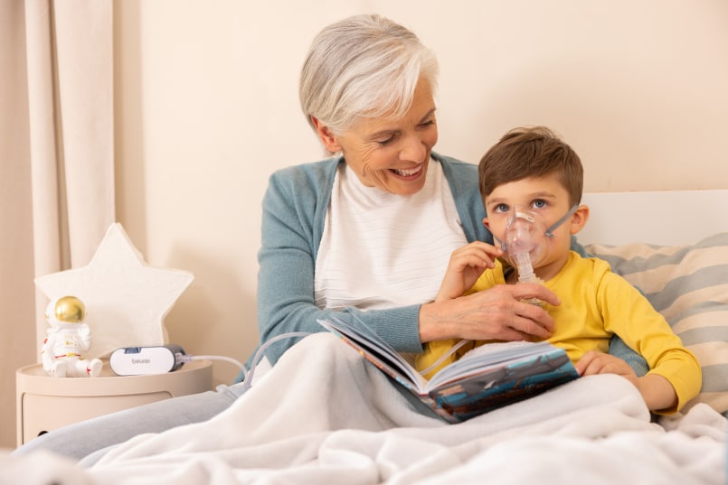 Ältere Frau sitzt mit einem Kind im Bett und hält ihm eine Inhalationsmaske ans Gesicht. Auf dem Schoß liegt ein geöffnetes Buch, daneben steht ein Inhalationsgerät.