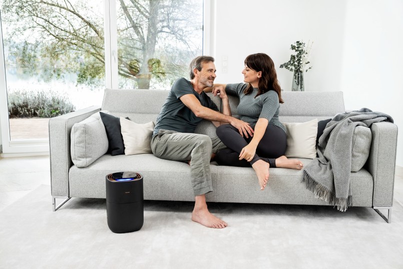 Ein Mann und eine Frau sitzen auf einem grauen Sofa und lächeln sich an. Der LB 300 steht links vom Sofa und leuchtet.