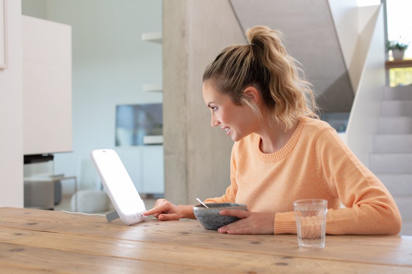 Jeune femme blonde assise à la table de salle à manger avec son petit-déjeuner, utilisant la lampe de luminothérapie TL 35