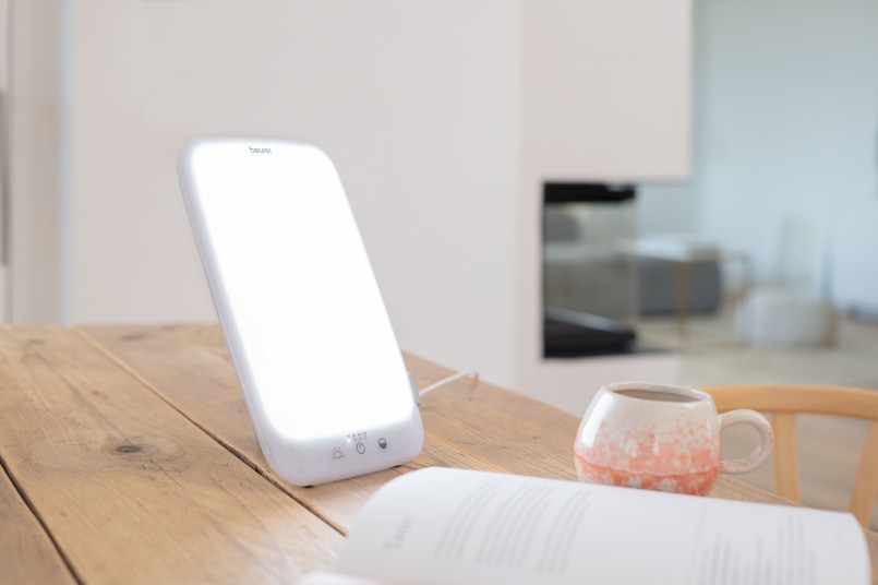 La lampe de luminothérapie TL 35 se trouve à côté d’un livre et d’une tasse sur une table en bois dans une salle à manger moderne