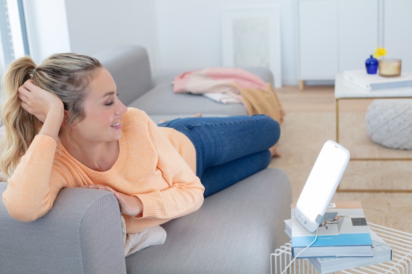 Jeune femme blonde allongée sur un canapé dans le salon, tandis que la lampe de luminothérapie TL 35 est dirigée vers son visage