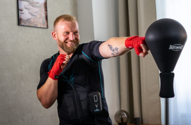 Christoph, l'entraîneur personnel, exécute un mouvement de kickboxing dans la combinaison EMS mobile d'Antelope. Il regarde un punching-ball en souriant. En arrière-plan, on devine un salon.
