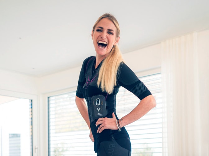 Presentatrice Anna Kraft is overtuigd door de EMS-training met Antelope. Ze draagt het mobiele EMS-pak van Antelope en kijkt met een glimlach in de camera. Ze draagt haar lange blonde haar in een paardenstaart en heeft haar handen langs haar zij geklemd. Op de achtergrond zijn ramen te zien.