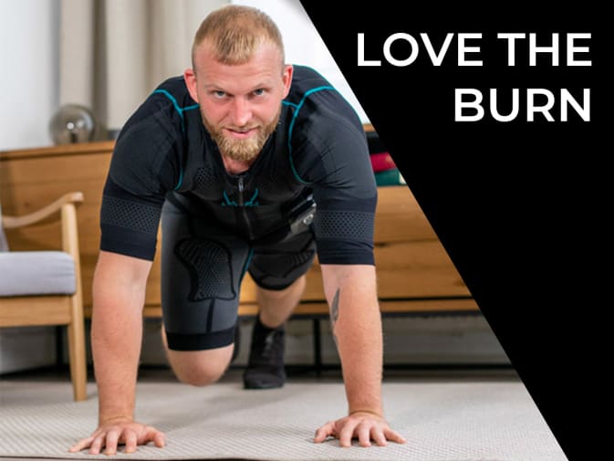 Personal trainer Christoph performs a fitness exercise on the floor with the mobile EMS suit from Antelope. He looks intently into the camera. Christoph has reddish-blonde hair and a reddish-blonde beard. A living room can be seen in the background.