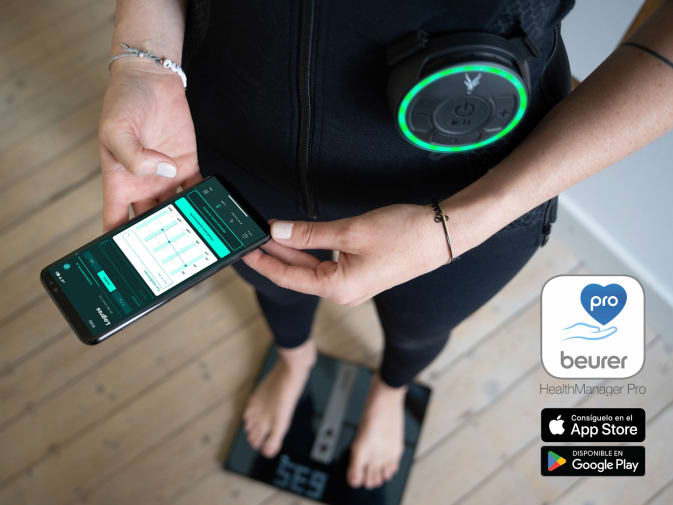 Una persona se pesa después de entrenarse con el traje EMS móvil de Antelope y hace un seguimiento de sus progresos en la app Antelope Go. La persona se ve hasta debajo del pecho y tiene las uñas pintadas de blanco. Alrededor de la persona se ve un cuarto de baño con suelo de madera.