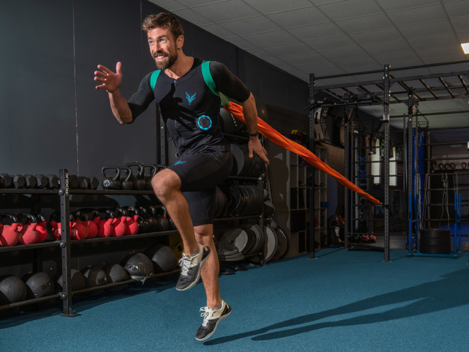 Ein sportlich gekleideter Mann in einem EMS-Anzug springt mit angelegtem Widerstandsband dynamisch nach vorne. Er trainiert in einem modernen Fitnessstudio mit Hanteln, Kettlebells und Trainingsgeräten im Hintergrund.