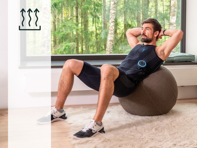 Mężczyzna rozgrzewa się przed treningiem w mobilnym kombinezonie EMS od Antelope. Siedzi na macie sportowej na tarasie w ogrodzie i rozciąga lewą nogę, uśmiechając się i patrząc w lewo. Ma brązową brodę i krótkie brązowe włosy.