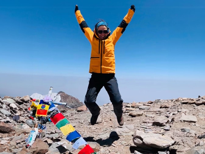 Anna Ott springt jubelnd mit erhobenen Armen auf einem felsigen Gipfel. Sie trägt eine orangefarbene Jacke und Sonnenbrille. Neben ihr flattern bunte Gebetsfahnen im Wind, im Hintergrund erstreckt sich ein klarer blauer Himmel.
