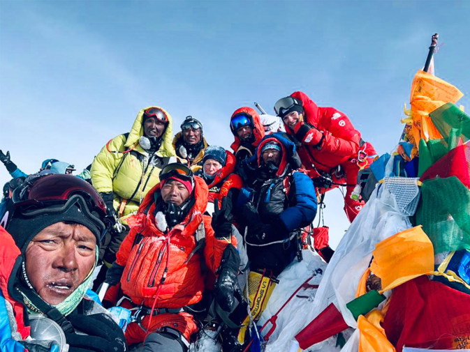 Eine Gruppe von Bergsteiger:innen in dicker Expeditionskleidung posiert auf dem Gipfel des Mount Everest vor klarem Himmel. Zwischen ihnen steht Anna Ott in roter Ausrüstung. Rechts im Bild wehen bunte Gebetsfahnen im Wind. Alle wirken erschöpft, aber stolz.