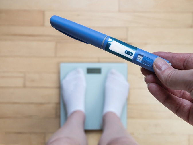 Eine Person wiegt sich auf einer Waage. Sie hält eine Abnehmspritze in der rechten Hand.