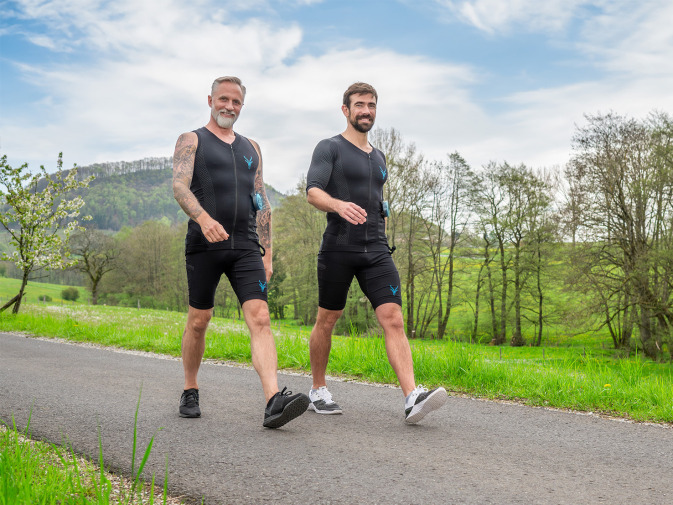 Zwei Männer im EMS-Anzug sind auf einem Spaziergang. Im Hintergrund ist eine grüne Landschaft zu sehen.