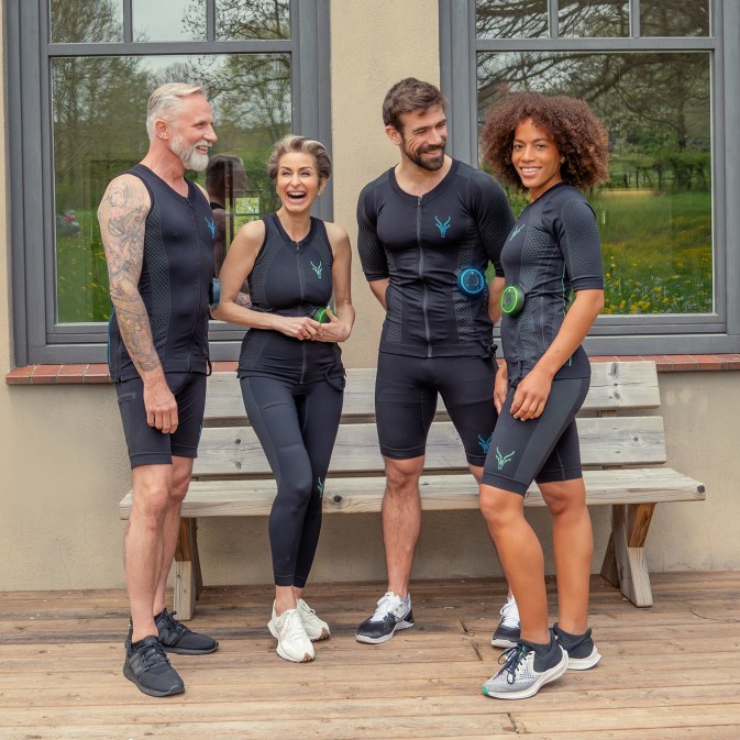 Eine Gruppe von sportlichen Personen steht vor einer Bank und lacht miteinander. Alle tragen einen EMS-Anzug.