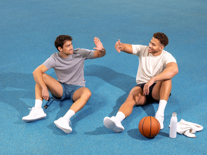 Zwei sportlich gekleidete Männer sitzen entspannt auf einem blauen Sportboden, geben sich im Sitzen ein High-Five und lächeln. Neben ihnen liegen ein Basketball, eine Trinkflasche, ein Handtuch und eine Massagepistole.