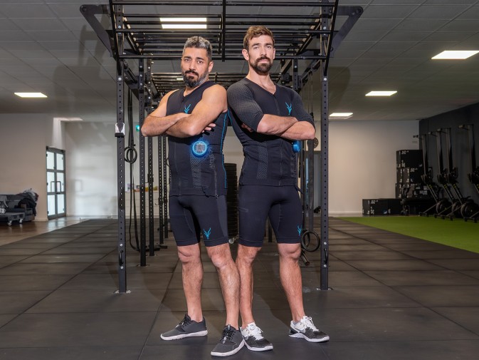 Zwei Männer stehen im Gym Rücken an Rücken. Der Mann links trägt das EMS Tank-Top von Antelope. Der Mann rechts den EMS Anzug.