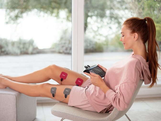 Młoda kobieta siedzi na krześle i przykłada do uda urządzenie EM 89 TENS/EMS firmy Beurer