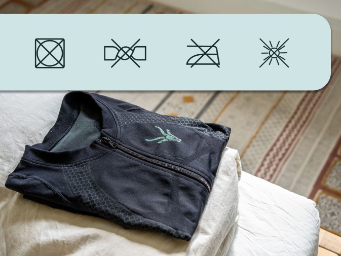 La combinaison EMS Antelope est posée pliée sur un lit. Au-dessus de l'image, les icônes « Ne pas sécher en sèche-linge », « Ne pas essorer », « Ne pas repasser » et « Ne pas exposer directement au soleil ».