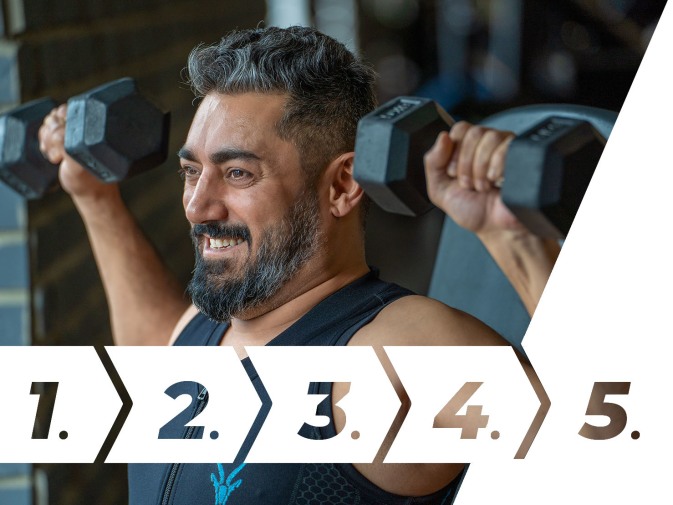 Ein Mann mit Bart trainiert im Fitnessstudio mit zwei Kurzhanteln und trägt ein schwarzes Sportshirt. Er lächelt konzentriert während der Schulterübung.