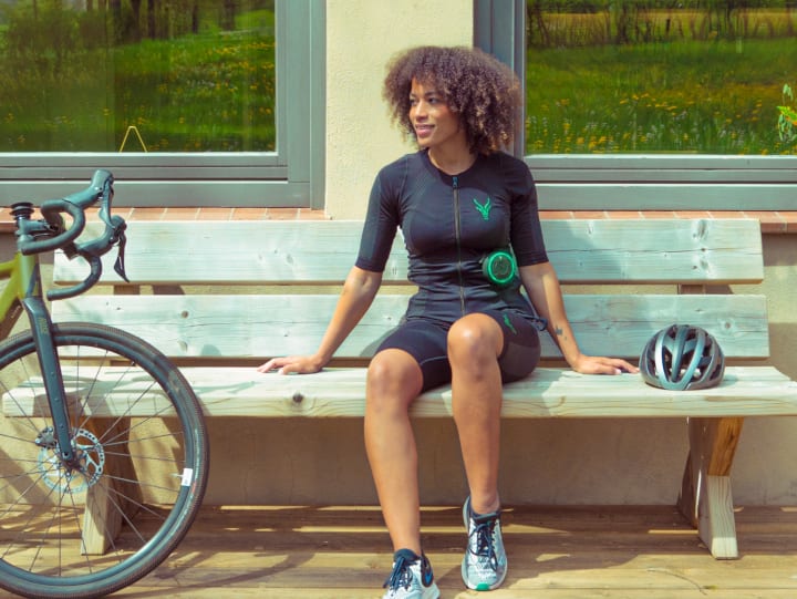 A woman is wearing an EMS suit while taking a break from cycling on her bike. She sits outside on a bench.