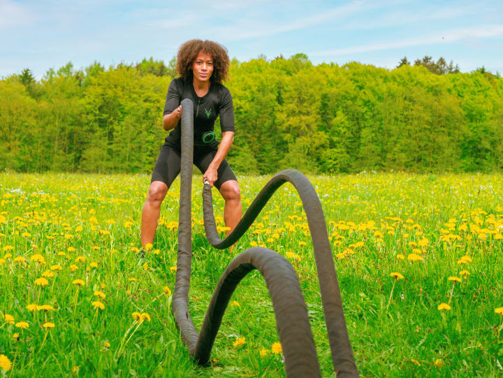 Frau trainiert im EMS-Anzug mit Battle Ropes auf einer blühenden Wiese vor Waldkulisse – funktionelles Outdoor-Training mit Elektrostimulation im Frühling.