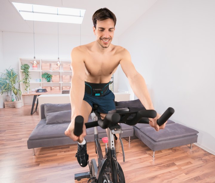 Un hombre hace un entrenamiento cardiovascular en casa en una bicicleta estática con el cinturón abdominal Corefit I EMS de Antelope.