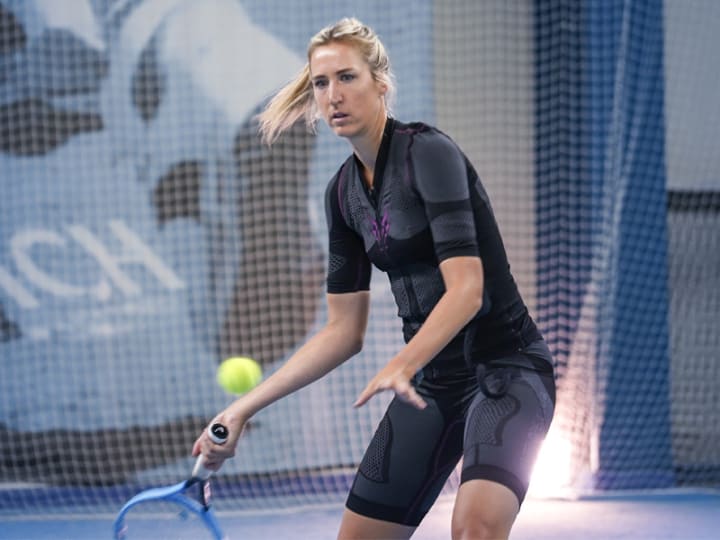 Sophia Bergner nutzt den EMS Anzug von Antelope für ihr Tennis-Training. Sie schaut lächelnd nach links. Ihre blonden Haare sind zu einem Pferdeschwanz gebunden. Im Hintergrund ist ein Netz zu sehen, das von der Decke hängt, sowie eine Wand in blauen Tönen.