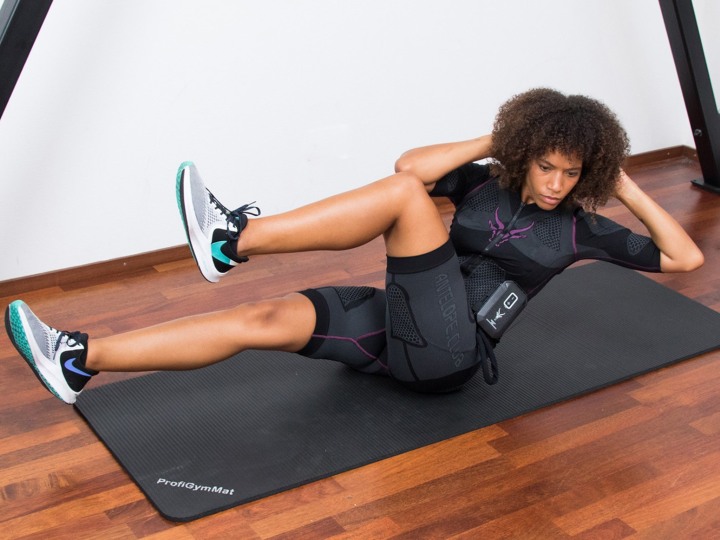 Eine junge Frau mit lockigen Haaren führt einen Sit-Up auf einer Sportmatte in dem EMS Anzug von Antelope durch. Sie hat ihr linkes Bein zu ihrem rechten geführt und schaut konzentriert.