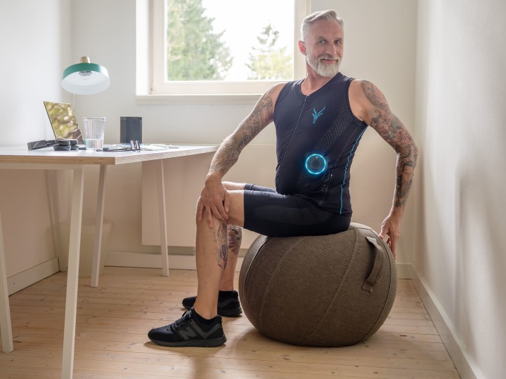 Älterer, tätowierter Mann trägt einen EMS-Anzug von Antelope und sitzt in einem modernen Homeoffice auf einem Sitzball, während er eine Trainings- oder Dehnübung macht.