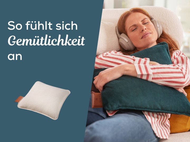 Junge Frau mit Kopfhörern kuschelt auf dem Sofa für gemütliche Wärme mit dem beurer Heizkissen HK77. Text "Made by us, loved by pets"
