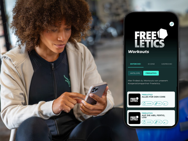 Eine Frau sitz im Gym mit Smartphone in der Hand. Rechts im Bild sieht man die Antelope Go App mit Freeletics Workouts.