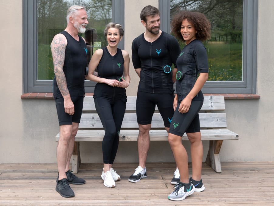 Vier Personen lachen miteinander, während sie den mobilen EMS Anzug von Antelope tragen. Die Personen schauen teilweise in die Kamera und teilweise schauen sie sich gegenseitig an. Im Hintergrund ist die Außenfassade von einem Haus erkennbar.