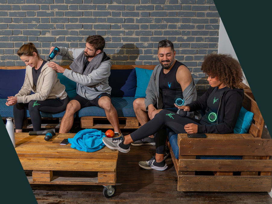 Four people sit on wooden benches in a cozy room with a brick wall. One woman receives a massage with a massage gun, while the others – two men and one woman in EMS training gear – sit relaxed, smile, or operate their device.