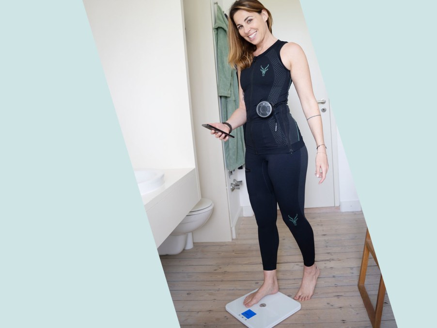 A woman wearing an EMS training suit stands barefoot on a body analysis scale in a bathroom, holding a smartphone. She is smiling at the camera.