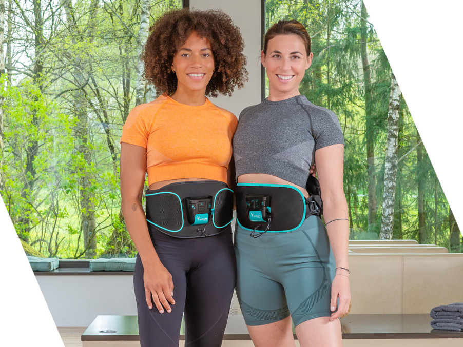 Two women stand side by side, each wearing an EMS belt around their waist. They are smiling at the camera in a bright room with large windows overlooking a green forest.