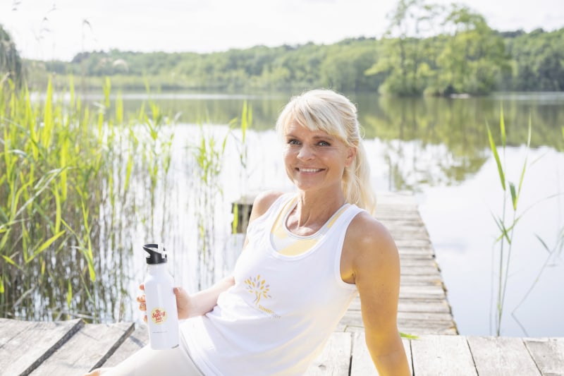 Carina Teutenberg sitzt auf einem Steg an einem See. Sie lächelt in die Kamera und hält eine Trinkflasche in der Hand.