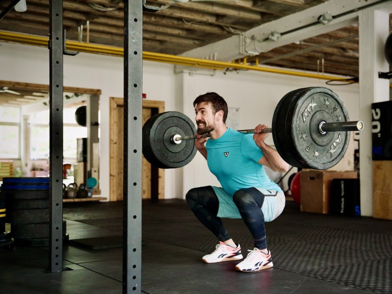 Olli Dehmel in einer tiefen Kniebeuge mit einer Langhantel mit Gewichten auf den Schultern. Er befindet sich in einem Fitnessstudio.