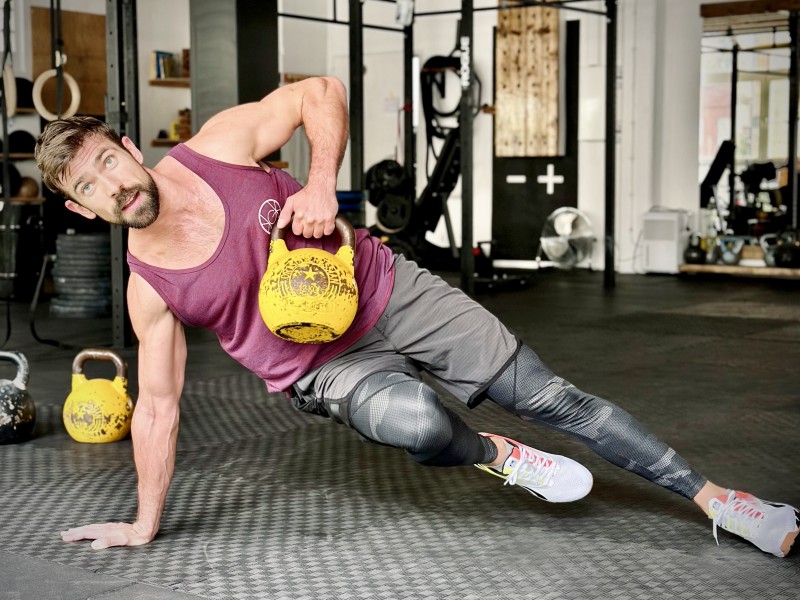 Olli Dehmel macht einen Side-Plank mit einer Kettlebell in einer Hand. Er befindet sich in einem Fitnessstudio.