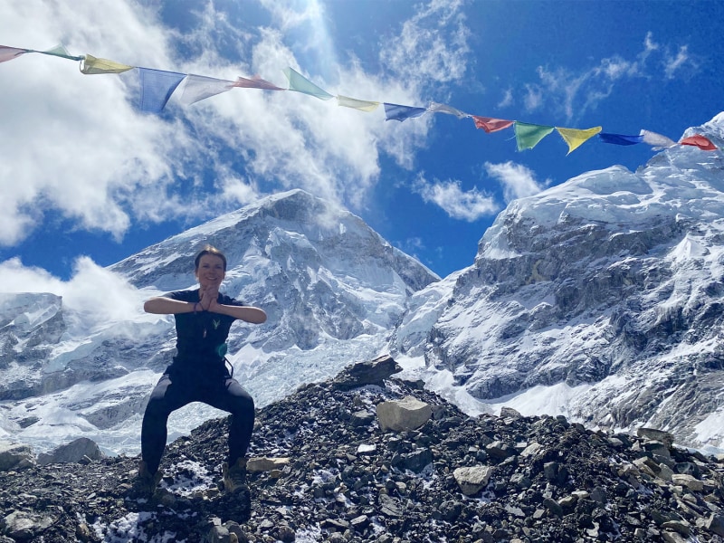 Anna Ott steht in einem EMS-Anzug auf felsigem Untergrund vor schneebedeckten Bergen und führt eine Yoga-Pose mit gefalteten Händen aus. Über ihr hängen bunte Gebetsfahnen im Wind.