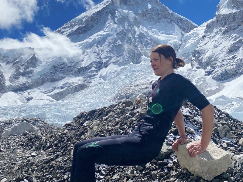Anna Ott sitzt in einem EMS-Anzug auf einem Felsen und stützt sich mit den Armen ab. Im Hintergrund ragt eine beeindruckende schneebedeckte Bergkulisse auf.