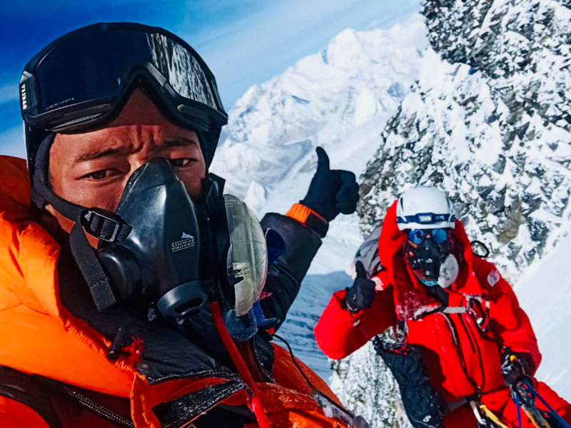 Im Vordergrund ein Bergsteiger in roter Expeditionskleidung mit Sauerstoffmaske. Im Hintergrund steht Anna Ott ebenfalls in rotem Anzug mit Steigeisen und Maske an einem schneebedeckten Hang. Beide zeigen den Daumen nach oben auf dem Weg zum Gipfel.