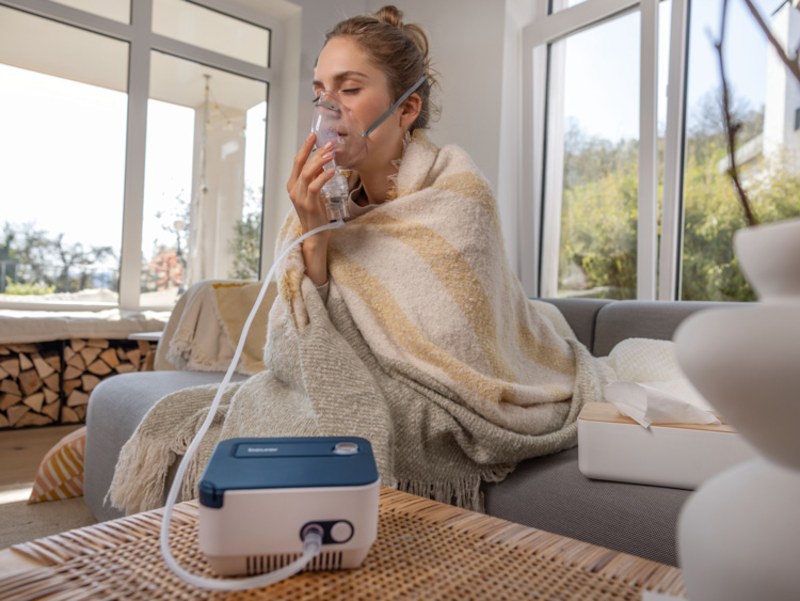 A woman is sitting on her sofa at home, using the Beurer IH 16 inhaler